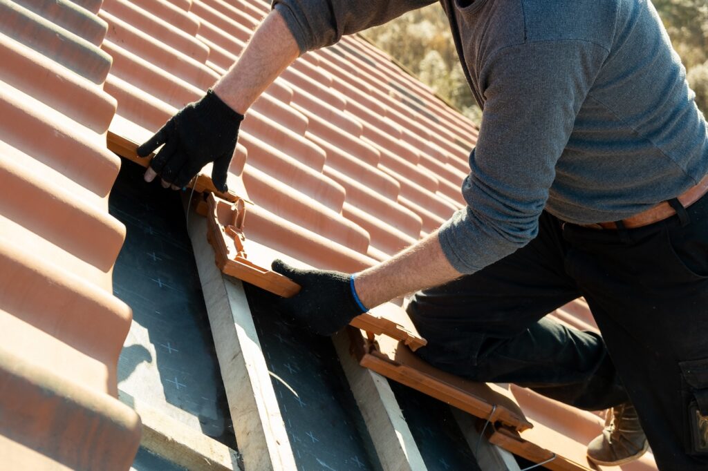 Closeup of worker hands installing yellow ceramic roofing tiles mounted on wooden boards covering residential building roof under construction.