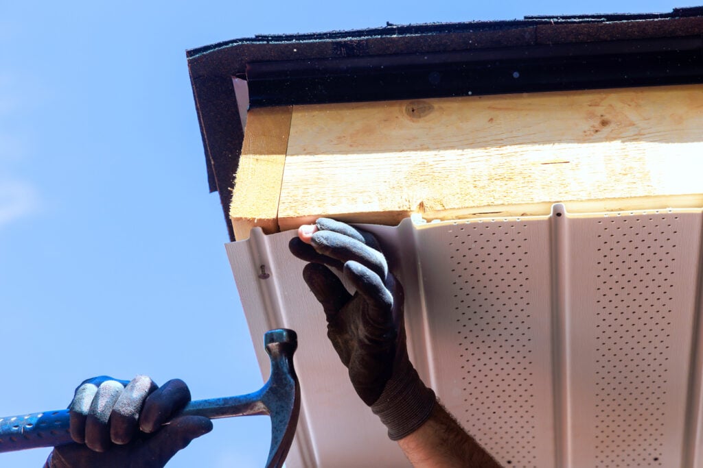 Contractor installs roof soffit panels using hammer wearing gloves under construction