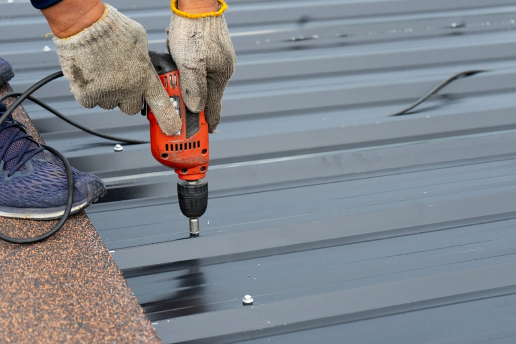 Workers installing the metal sheet roof by electrical drilling machine. Selective focus on the drilling tool while building the roof