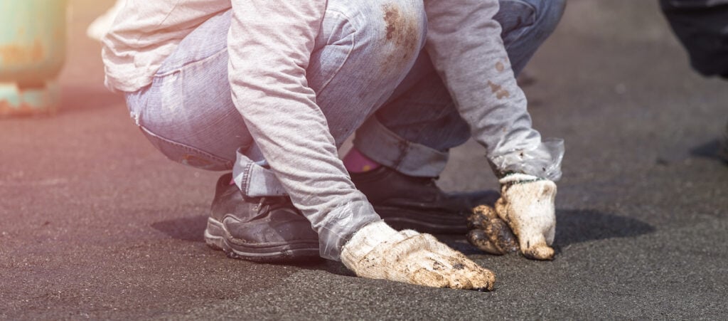 Worker applying black roofing sealant on a flat roof surface with gloved hands