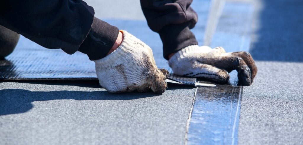Worker sealing a seam on flat roofing membrane with gloved hands and a hand tool